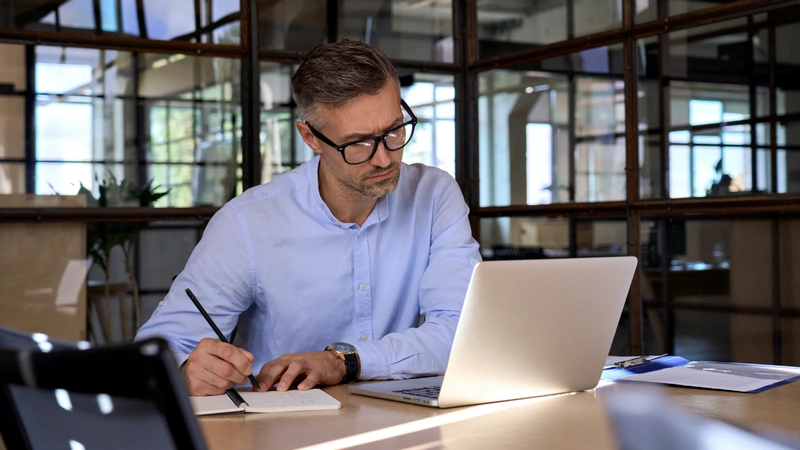 A man sat at a table making notes from content on his laptop screen